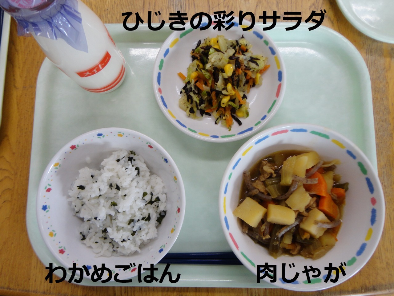 手前にわかめごはんと肉じゃが、奥に牛乳とひじきの彩りサラダを並べた給食の写真