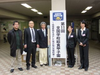 会場の案内板の前で関係者5名が記念撮影をしている写真