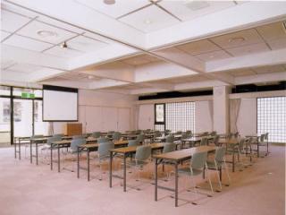 明るい光が差し込んでおり、正面にスクリーン、二人掛けの長机と椅子が並んでいる研修室1の室内写真