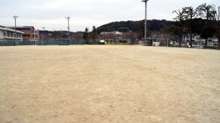 フェンスの奥に建物や小高い山があるグラウンドの写真