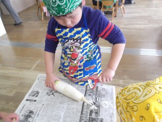 三角巾とエプロンを身に着けた男の子が新聞紙の上で大根の皮をビューラーを使ってむいている写真