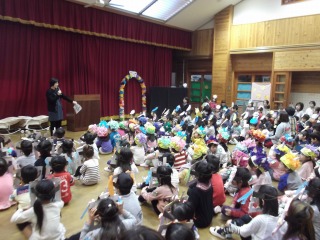 前方の舞台の前に一人の先生が話をしており、園児の皆さんは座って節分集会に楽しく参加している様子の写真