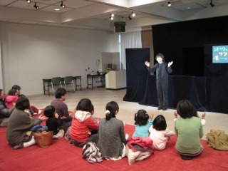 黒い布が掛けられた台の前で全身黒い服を着た人形劇の方が話をしており、親子の参加者の皆さんが話を聞いている写真
