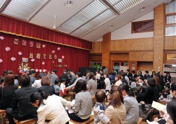 舞台の幕に「ごにゅうしょ おめでとう」と掲示されており、ホールに集まった保護者と子どもが一緒に入園式に参加している写真