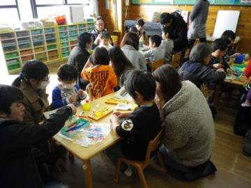 鬼のお面づくりをしている子供たちの横に座り、手元を見ている保護者や家族の写真