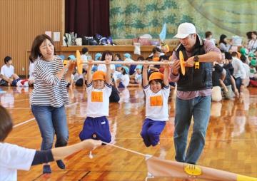 バーベルにぶら下がる園児2名を、男女が持ち上げ運んでいる写真