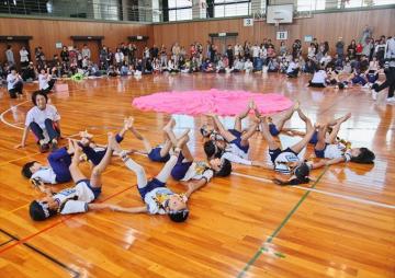 手をつないで床にあおむけの園児が、両足を広げ隣の園児と足をくっつける組体操をしている写真
