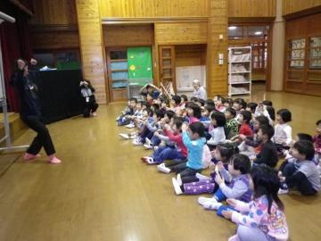 前方に立ち、踊るあかいとみこ先生と、座って両手で丸のポーズをする園児の写真