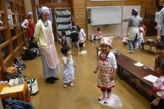 三角布とエプロンを身に着けた子どもが袋に入ったうどんを足で踏んでおり、その様子を笑顔で見守っている母親の写真
