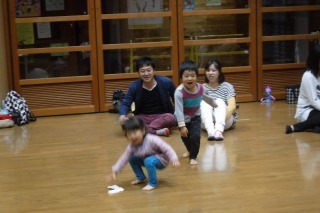 男の子と女の子が競って前方に落ちている靴下を走って拾おうとしており、その様子を後ろに座っているパパとママが見守っている写真
