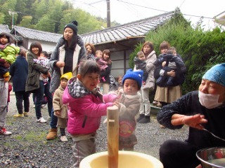 こども2人でおもちつきをしているのを見ている親子の写真