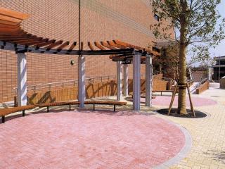 丸い形に装飾されたアスファルトで憩いのスペースが設けられ、ベンチと上部には木材を扇形に均等に並べ日よけが設置されている触れ合い広場の写真