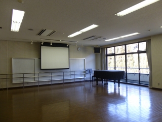 正面にスクリーン、右側に大きな窓、右前にグランドピアノが置かれ、床がフローリングの研修室2の室内写真