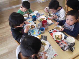 机にご飯やおかずの入ったお皿、豚汁が並んでおり、子ども達が美味しそうに味わって食べている写真