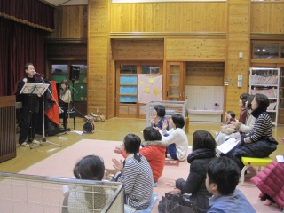 譜面台の前で歌を歌っている古川先生と手拍子をしながら楽しんでいる参加者の皆さんの写真