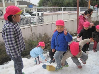 ピンク色の帽子を被った園児が外に積もった雪の上で雪遊びをしている写真