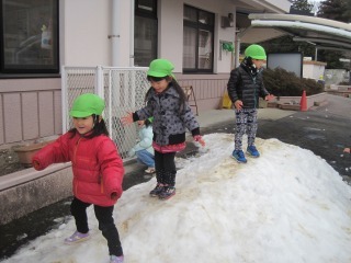 雪で出来た山の上から立ったまま少しずつ滑る遊びをしている緑色の帽子を被った3人の園児の雪遊びの写真