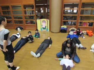 参加者の方々が床に仰向けに寝ており、バレエ講師の高山さんが参加者の皆さんの前に立って指導をしている写真