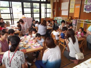 子ども達がグループごとに分かれて机に座って給食を食べており、保護者が子どもの側で、給食を食べている様子を見ている給食参観の様子の写真