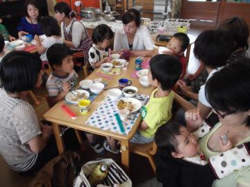 子ども達がテーブルの席に着き、給食を食べており、保護者が子どもの側で給食を食べているのを見ている給食参観の様子の写真