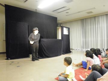 黒い布が掛けられた舞台の横に黒い服を着た男性が立って子ども達に話をしている人形劇の様子の写真