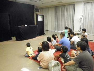 黒い幕が掛けられた大きなセットの前に、参加者親子が座っており、人形劇を見ている写真