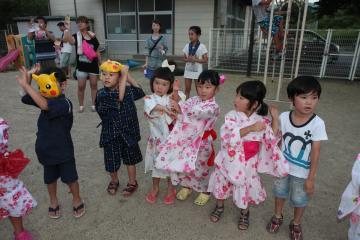 キャラクターのお面を被った男の子や浴衣を着た女の子達が園庭で両手を顔の横に挙げて踊っている写真