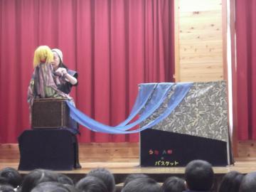舞台上で人形を操るやおちあきさんと人形劇を見ている子どもたちの写真