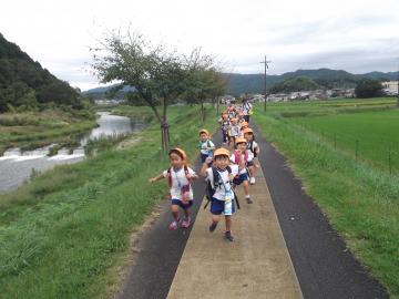 リュックサックを背負い、水筒を肩に下げた3歳児が土手を走っている写真