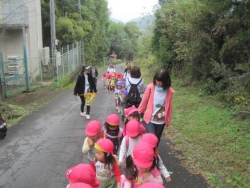 坂道で、列になる園児と、その横について歩く職員と一緒にお散歩している写真