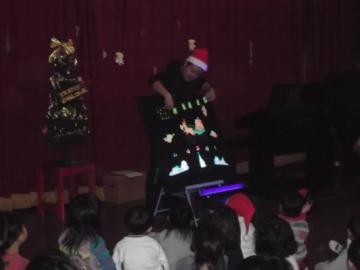 サンタの帽子をかぶった男性が、暗闇で光るパネルシアターを上演し、注目している園児の後ろ姿の写真