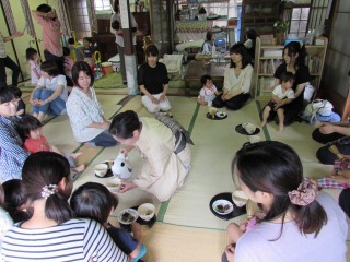 ござの上に親子が座り、着物を着た女性がお椀にお湯を注いでいる写真