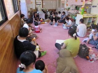 ピンクのカーペットの上で親子が円になって座り、べビーマッサージの話を聞いている写真