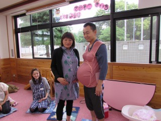おなかの大きな母親と、妊娠シミュレーターを着用した父親が立っている写真
