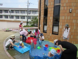 タイルの建物の前の屋外に置かれた3つのプールでママと子どもたちが、水あそびをしている写真