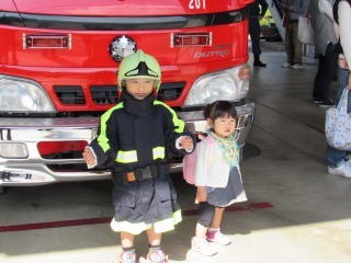 赤い消防車の前に立つ、幼児用消防士の服を着ている男の子の写真