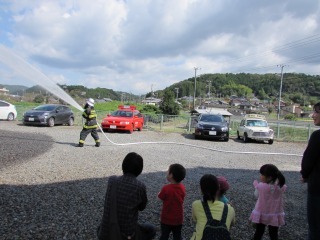 消防服を着た消防士さんがホースを持って構え、放水が行われており、その様子を参加者の親子がみている写真