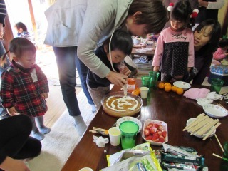 ホイップクリームを絞ってケーキを作っている男の子と後ろからサポートしている母親の写真