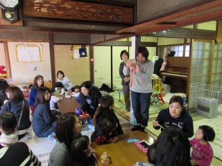参加者親子がテーブルごとに分かれて座っており、中央に立つ女性の方をみながら、わらべうたあそびをしている写真