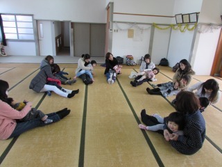 畳の部屋で参加者が輪になって、小さな子どもを膝の上に座らせて、ふれ合いあそびをしている写真